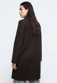 Hnědý kabát z vlněné směsi s přizpůsobeným střihem, dlouhými rukávy a klopovým límcem, doplněný světle modrými džínami. Hladká textura.