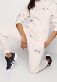 Beige bomullshuvtröja och sweatpants-set med en logotyp på bröstet. Koordinerat med svarta träningsskor. Slät textur, avslappnad design.