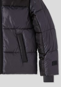 Czarna, pikowana kurtka z zamkiem błyskawicznym, z kieszenią, ściągaczem na mankiecie i naszywką z logo TRN na rękawie, na jednolitym tle.