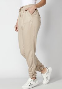Personne portant un pantalon de jogging beige avec cordon et poignets élastiques ainsi que des baskets blanches avec des motifs d'étoiles argentées, debout devant un fond uni.