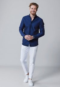 Donkerblauwe overhemd met lange mouwen, gecombineerd met witte slim-fit broeken en witte sneakers, staande op een neutrale achtergrond.