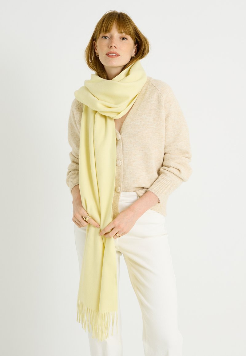 Bufanda suave amarilla con bordes de flecos, hecha de tela ligera, colocada alrededor del cuello, combinada con un cárdigan beige y pantalones blancos.