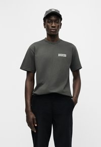 Νεαρός άνδρας που φοράει σκούρο γκρι μπλουζάκι Dickies και αντίστοιχο καπέλο, στέκεται με το ένα χέρι στην τσέπη μπροστά σε απλό λευκό φόντο.