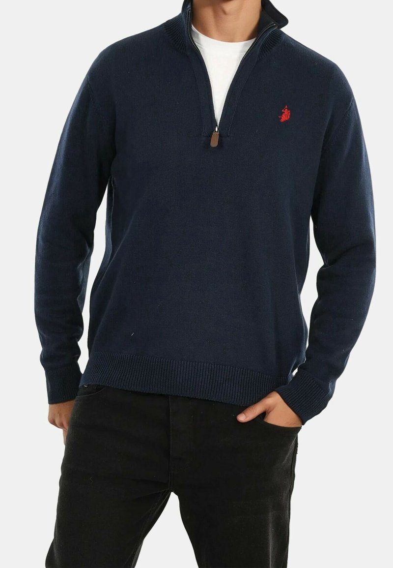 Homme portant un pull à col zippé bleu marine avec un logo rouge sur la poitrine, un sous-vêtement blanc et un pantalon noir, la main dans la poche.