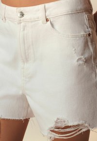 Biele denimové šortky majú zapínanie na gombík a zips, poškodené detaily, rozstrapkané okraje a vrecká vpredu aj vzadu.
