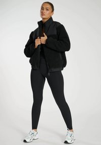 Schwarze Fleece-Bomberjacke mit einer glatten, gepolsterten Vorderseite, Reißverschluss und gerippten Bündchen. Getragen über schwarzen Leggings und sportlichen Sneakers.