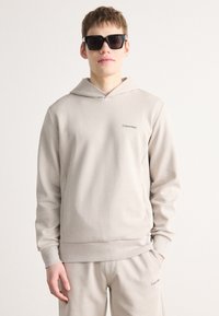 Béžová mikina s kapucňou s hladkým povrchom, na hrudi s malým logom Calvin Klein. Doplněná tmavými slnečnými okuliarmi.