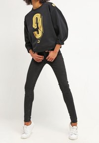 Sweat-shirt noir avec numéro graphique doré "9" sur le devant, associé à un jean skinny noir et des baskets blanches. Le sweat-shirt présente des manches surdimensionnées.