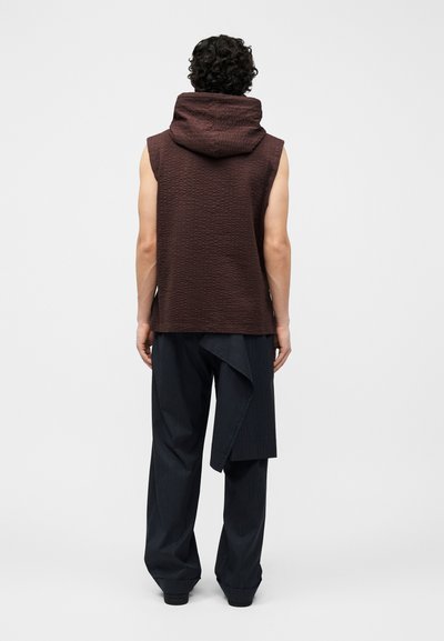 Man met krullend haar, gekleed in een mouwloos bruin met capuchon en zwarte broek met fijne strepen en asymmetrische details, staande met de rug naar voren tegen een witte achtergrond.