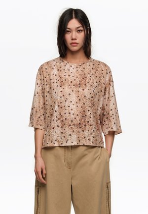 Žena oblečená v béžovom, voľnom topu s hnedo-čiernym škvrnitým vzorom a béžových nohaviciach typu cargo s bočnými vreckami na bielom pozadí.