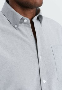 Harmaa nappikauluspaita, jossa on teksturoitu kangas, klassinen kaulus, valkoiset napit ja yksi rintatasku.