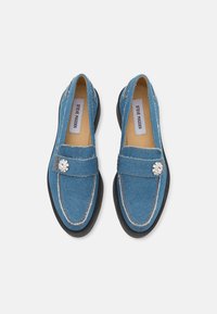 Blå denim loafers med sort gummisål, der har en dekorativ rhinestone-accent og kontrasterende syninger.