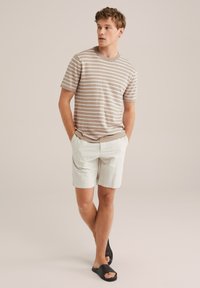 Tricou stripe bej și alb combinat cu pantaloni scurți bej deschis. Modelul poartă sandale negre, stând cu mâinile în buzunare.
