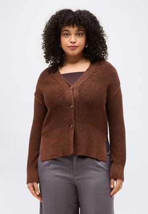 Keharapärase soenguga naine, kellel on pruunid nööbitavad kootud kardigan, tume pluus ja hallid püksid, seisab lihtsa heleda tausta ees.