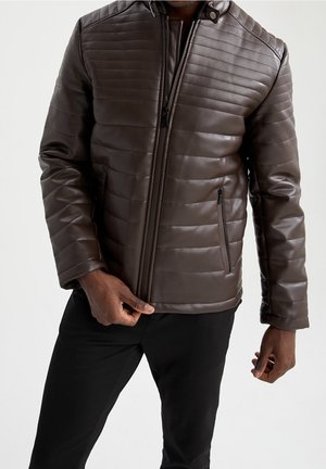 Homme portant une veste en cuir matelassée marron foncé avec poches zippées et pantalon noir sur un fond clair.