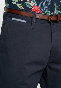 Pantalon en coton bleu marine à la texture lisse, avec un accent en denim sur la poche et une ceinture en cuir marron.