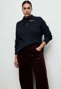 Jersey de punto azul oscuro con cuello alto y tres botones dorados, combinado con pantalones de pana burdeos de pierna ancha. Textura suave y ajuste relajado.