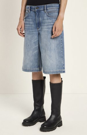 Person wearing knee-length light blue denim shorts and black mid-calf leather boots with thick soles, standing against a plain light background.