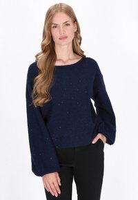 Vrouw met lang golvend blond haar, draagt zilveren oorringen, een donkerblauwe gespikkelde trui en zwarte broek, poseert met één hand in de zak.