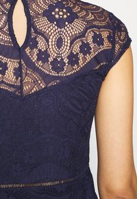 Tmavě modré krajkové šaty s květinovým vzorem a texturovanou látkou, s vykrojeným detailem na zádech a krátkými rukávy.