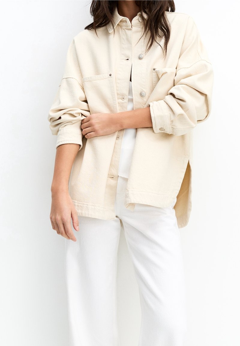 Femme en chemise beige oversize boutonnée avec manches retroussées et pantalon blanc, debout devant un fond blanc.