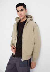 Veste bomber beige à capuche avec poches avant, fermeture éclair et poignets côtelés. Portée par-dessus une chemise marron foncé et un pantalon cargo gris.
