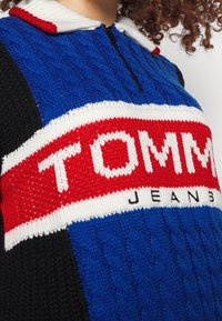 Maglione a maglia con zip a un quarto, caratterizzato da una combinazione di colori blu e nero, colletto bianco e un logo "TOMMY JEANS" rosso e bianco ben visibile.