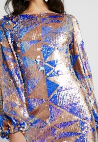 Paljetklänning med geometriska mönster i blå, guld och kopparfärger; långa ärmar med samlade manschetter; slät yta och reflekterande yta.