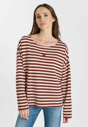 Donna con lunghi capelli castani che indossa un maglione largo a righe bordeaux e beige e jeans blu su uno sfondo chiaro e uniforme.