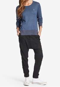 Blauer Strickpullover mit geripptem Saum und Bündchen, kombiniert mit lockeren schwarzen Hosen und weißen High-Top-Sneakern. Mit kleinem Logo auf dem Pullover.