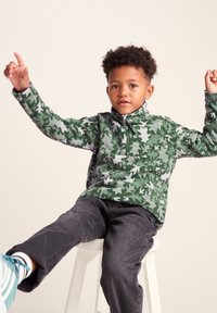 Green fleece pullover with a leaf camouflage pattern, half-zip collar, and long sleeves, paired with dark gray pants and teal-accented shoes.