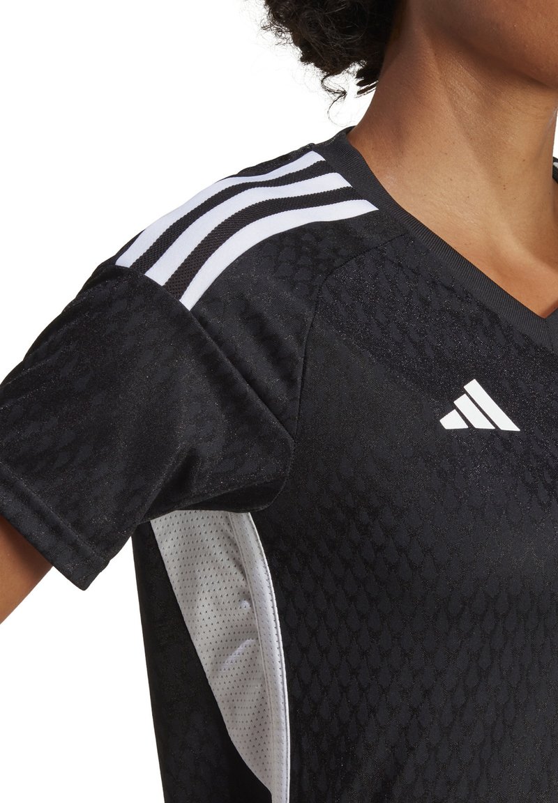 Czarny sportowy jersey z teksturowanej tkaniny, białe siateczkowe panele boczne i trzy białe paski na ramieniu. Posiada dekolt w kształcie litery V.