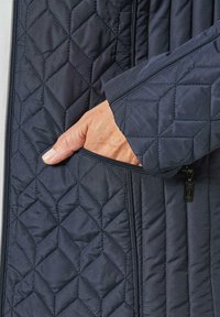 Marineblaue gesteppte Jacke mit einem strukturierten Rautenmuster, seitlichem Reißverschlussfach und einer glatten Stoffoberfläche. Eine Hand ist sichtbar, die in die Tasche greift.