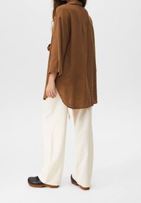 Femme portant une chemise longue marron ample, un pantalon large crème et des sabots noirs à semelles en bois, se tenant contre un fond uni.