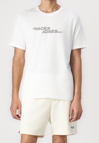 Weißes Baumwoll-T-Shirt mit Rundhalsausschnitt, versehen mit einem grauen Grafikdruck, der "JACK & JONES" liest. Kombiniert mit hellbeigen Shorts.