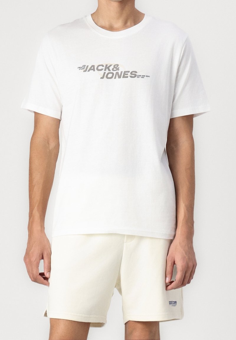 Weißes Baumwoll-T-Shirt mit Rundhalsausschnitt, versehen mit einem grauen Grafikdruck, der "JACK & JONES" liest. Kombiniert mit hellbeigen Shorts.