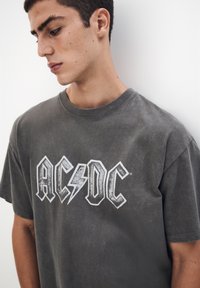 Siva bombažna majica z udobnim izrezom in kratkimi rokavi, na kateri je uveljavljen logotip AC/DC v beli barvi z raketo.