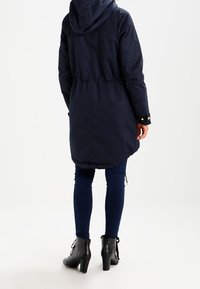 Chaqueta larga azul marino con capucha y cintura ajustada. Cuenta con elástico en el dobladillo y botones a presión en los puños. Combinada con jeans azul oscuro y botas negras hasta el tobillo.