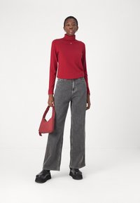 Maglione rosso a collo alto, jeans grigi a gamba larga, scarpe con piattaforma nere e una piccola borsa a tracolla rossa. Il modello si trova di fronte a uno sfondo neutro.