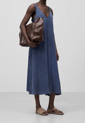 Person trägt ein ärmelloses blaues Midikleid, hält eine große braune Ledertasche und trägt beige Slidesandalen vor einem einfarbigen Hintergrund.