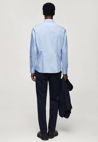 Uomo in piedi di spalle, con una camicia a maniche lunghe azzurro chiaro, pantaloni scuri, scarpe nere e che tiene un giubbino scuro con la mano destra.