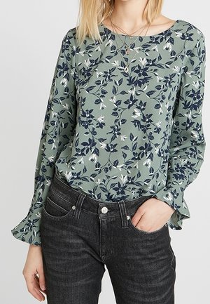 Femme portant un chemisier vert à fleurs à manches longues avec des poignets volantés, un jean taille haute sombre, une main dans la poche et des colliers superposés.