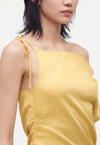 Femme aux cheveux noirs mi-longs portant un top jaune en satin sans manches avec une bretelle nouée sur fond blanc.