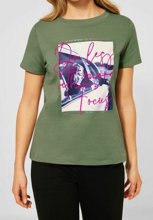 Vrouw draagt een olijfgroen T-shirt met een zwart-wit foto en roze tekst op de voorkant met de woorden "Doe minder met meer focus".