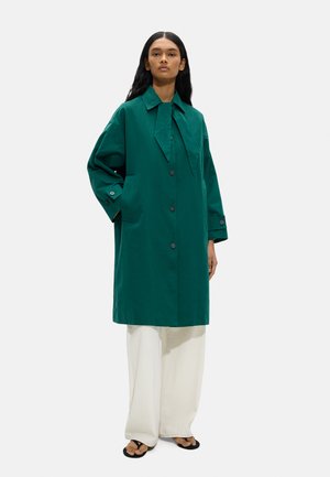 Kobieta ubrana w długi, ciemnozielony płaszcz na guziki z detalem krawata, białe spodnie o szerokich nogawkach i czarne sandały, stojąca na jednolitym tle.