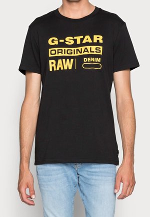 Mężczyzna ubrany w czarną koszulkę z żółtym napisem „G-STAR ORIGINALS RAW DENIM” oraz jasnoniebieskie jeansy, stojący na jednolitym tle.