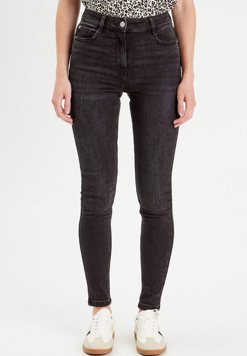Jean skinny noir taille haute avec une texture en denim lisse, design à cinq poches et passants de ceinture, associés à des baskets blanches.
