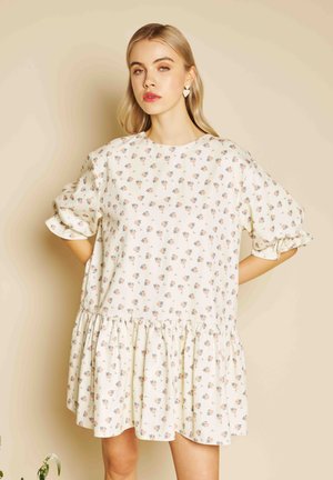 Jauna moteris dėvi laisvą baltą suknelę su mažais gėlių raštais ir rauktomis briaunomis, stovinti priešais smėlio spalvos foną.