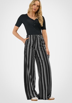 Kobieta stoi, ubrana w czarną koszulkę z krótkim rękawem i szerokie czarne spodnie w białe pionowe paski, ręce w kieszeniach, neutralny wyraz twarzy.