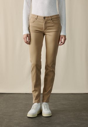 Kvinde iført beige slim-fit bukser, hvid langærmet top og hvide sneakers, står på gråt gulv med beige baggrund.